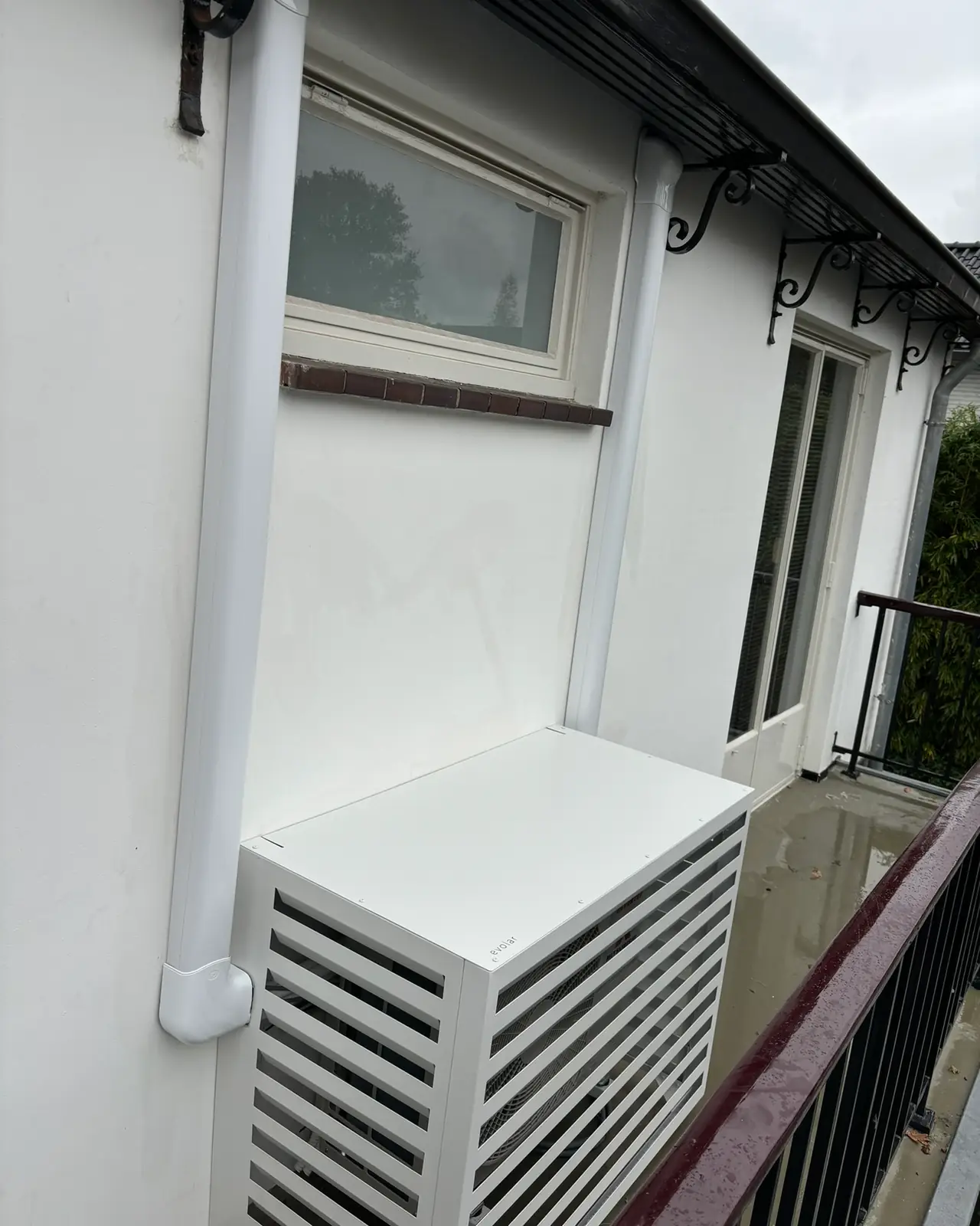 Een buitendeel in een witte lamellenkast op een balkon; de leiding loopt in een witte goot langs de gevel.