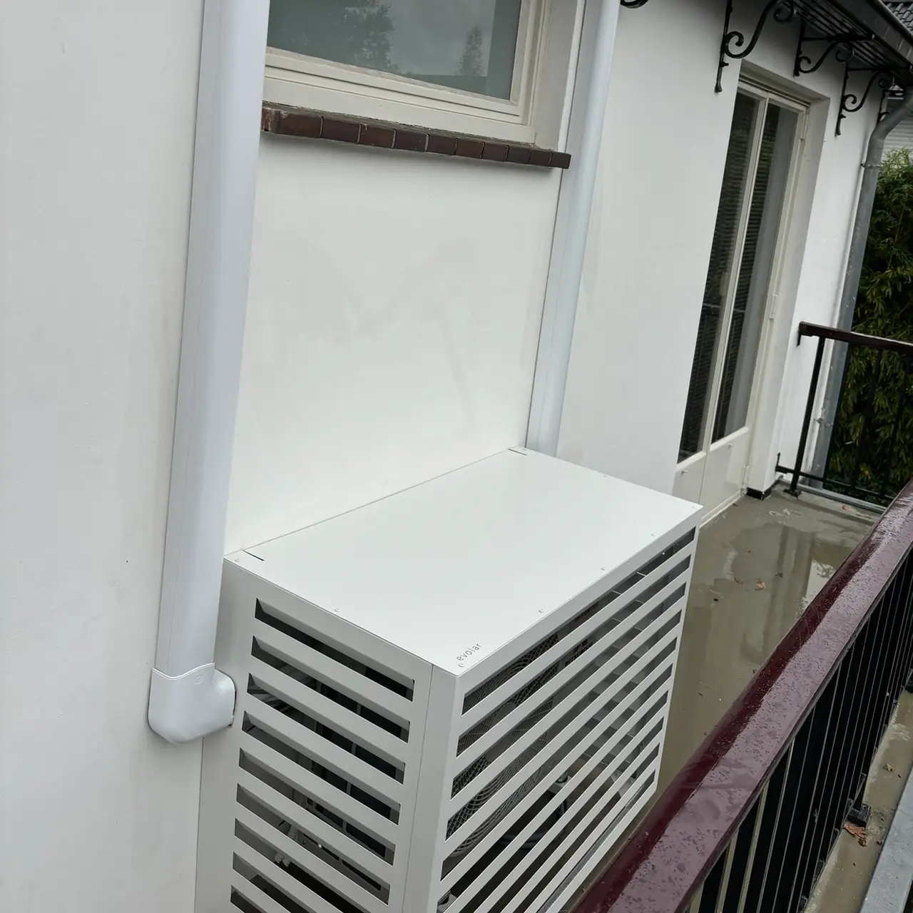 Een buitenunit in een witte omkasting op een balkon, met de leiding in een witte goot langs de gevel.