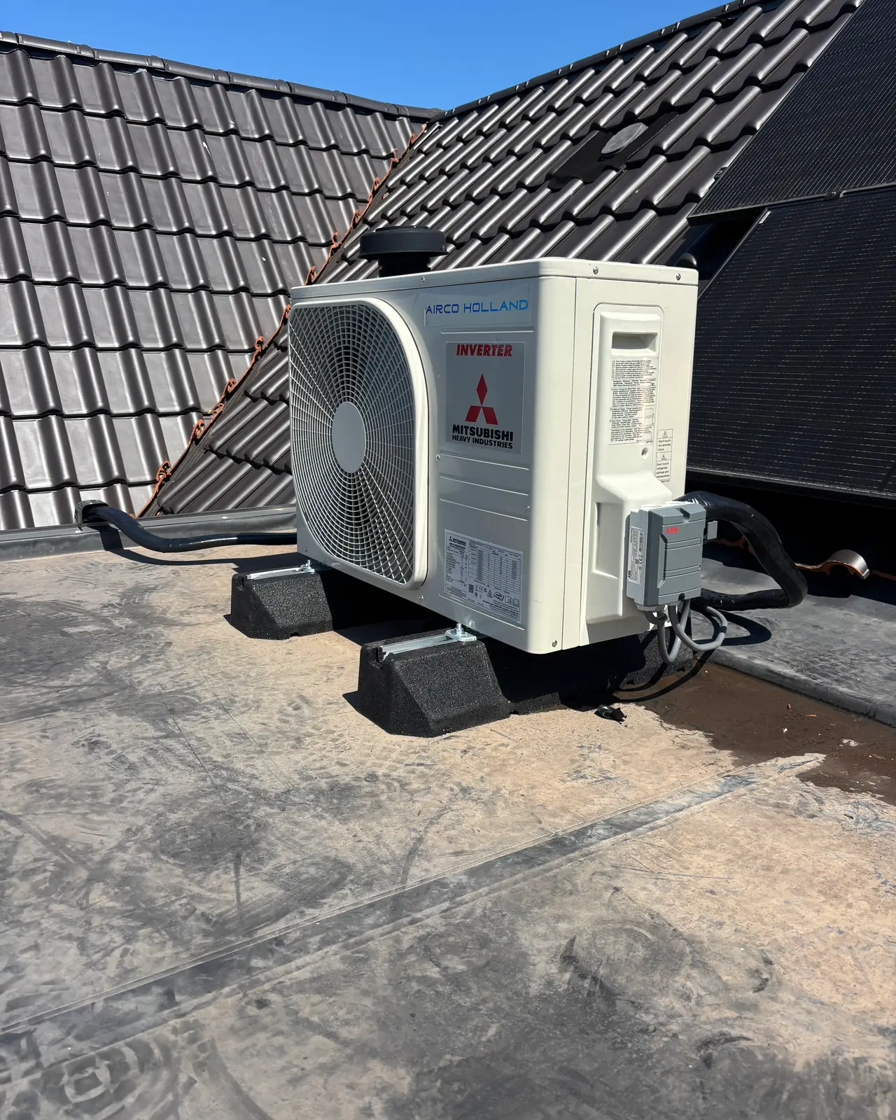 Een nieuwe buitenunit van Mitsubishi Heavy Industries op rubberen voeten op een plat dak, met een pannendak erachter.