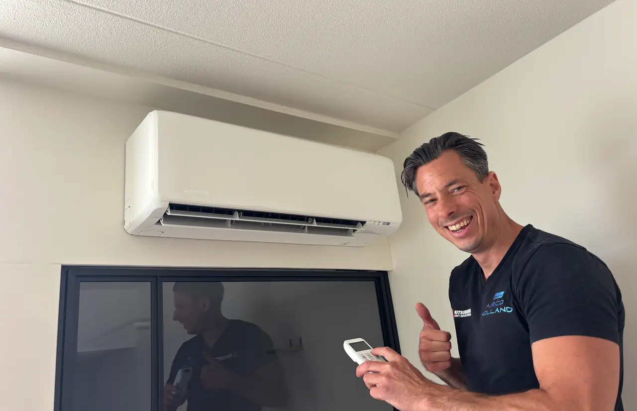 Peter lacht met de afstandsbediening in zijn hand naast een nieuw wit wandmodel van Mitsubishi Heavy Industries.