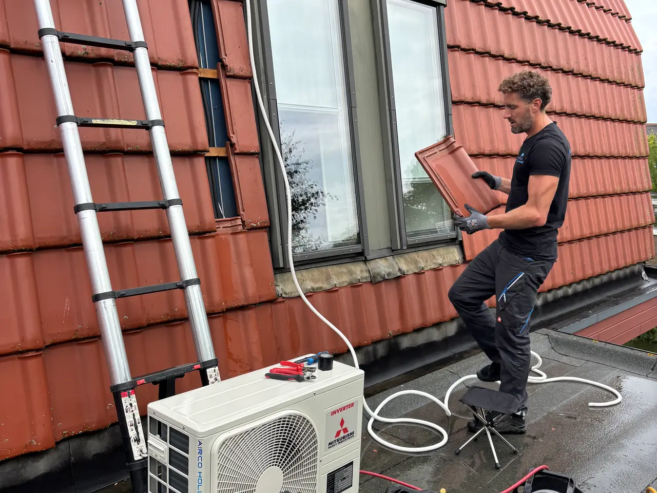 Rein houdt een losse dakpan vast bij een raam in een pannengevel; een witte leiding loopt naar het buitendeel.
