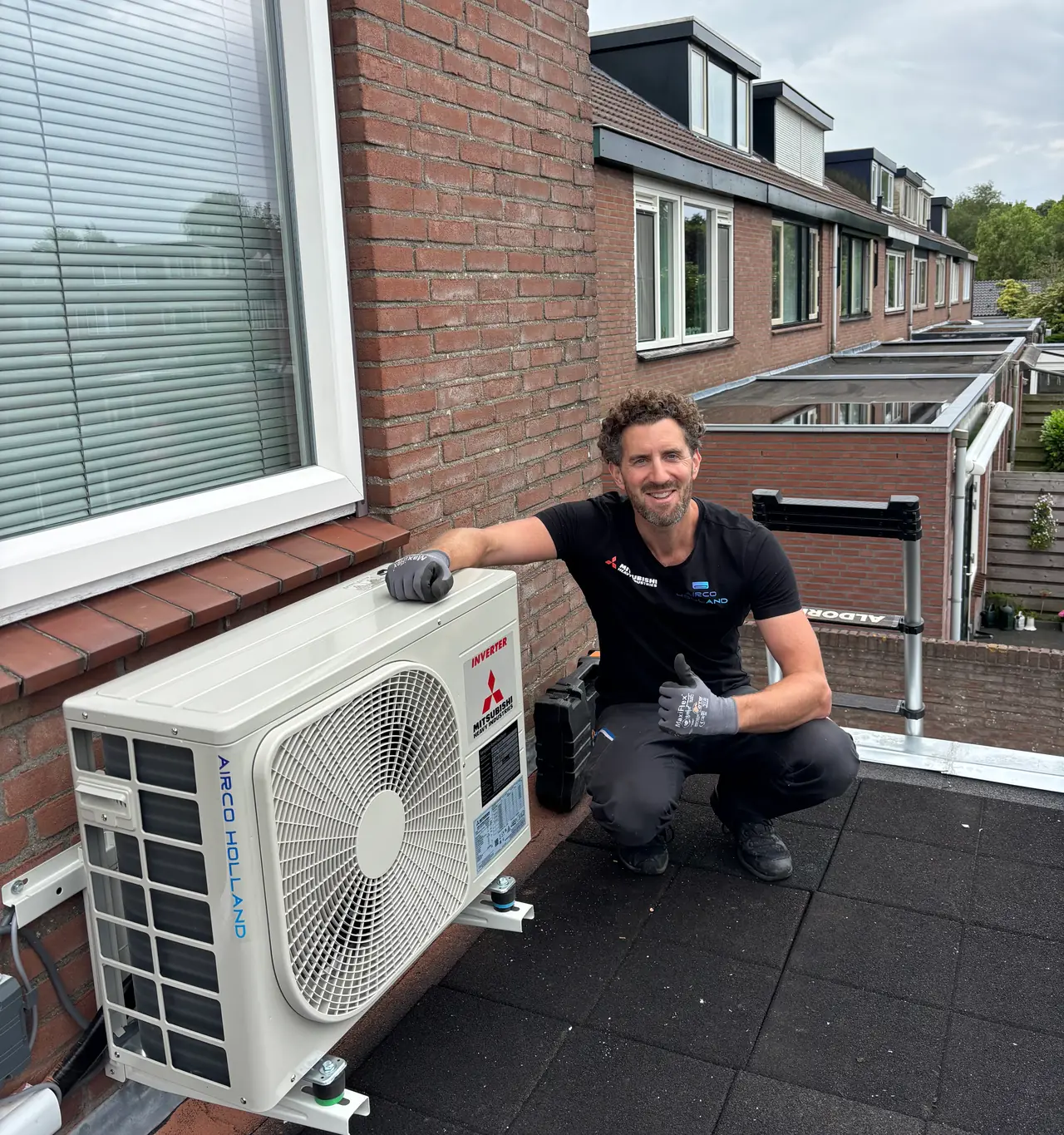 Rein van Airco Holland hurkt lachend naast een nieuw buitendeel op een dakterras, met een rij huizen erachter.